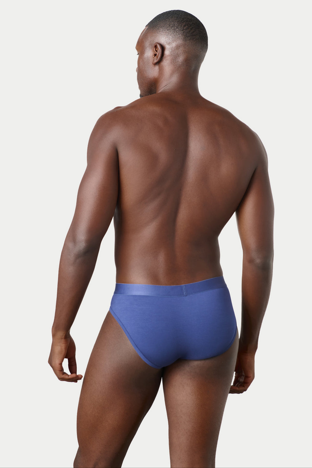A man shown from the back wearing a pair of blue briefs against a light gray studio background.