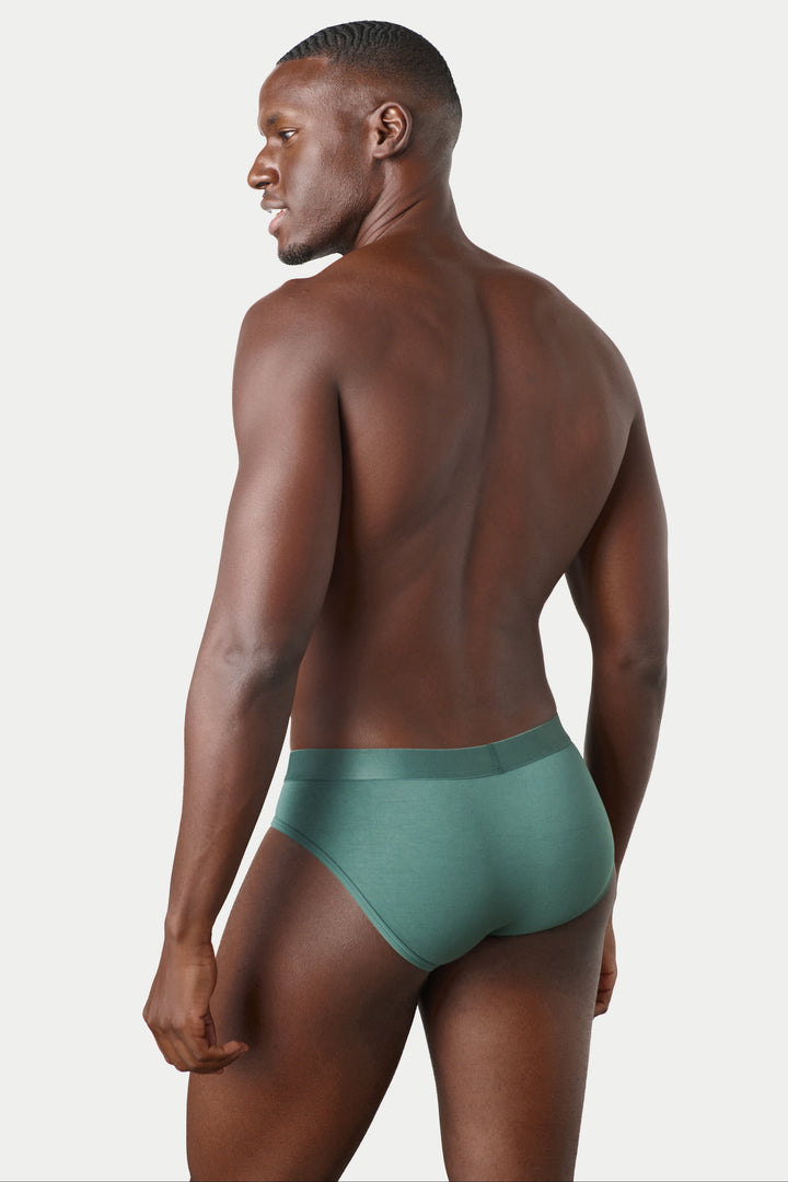 A man shown from the back wearing a pair of low-rise, solid teal-green briefs against a light gray studio background.