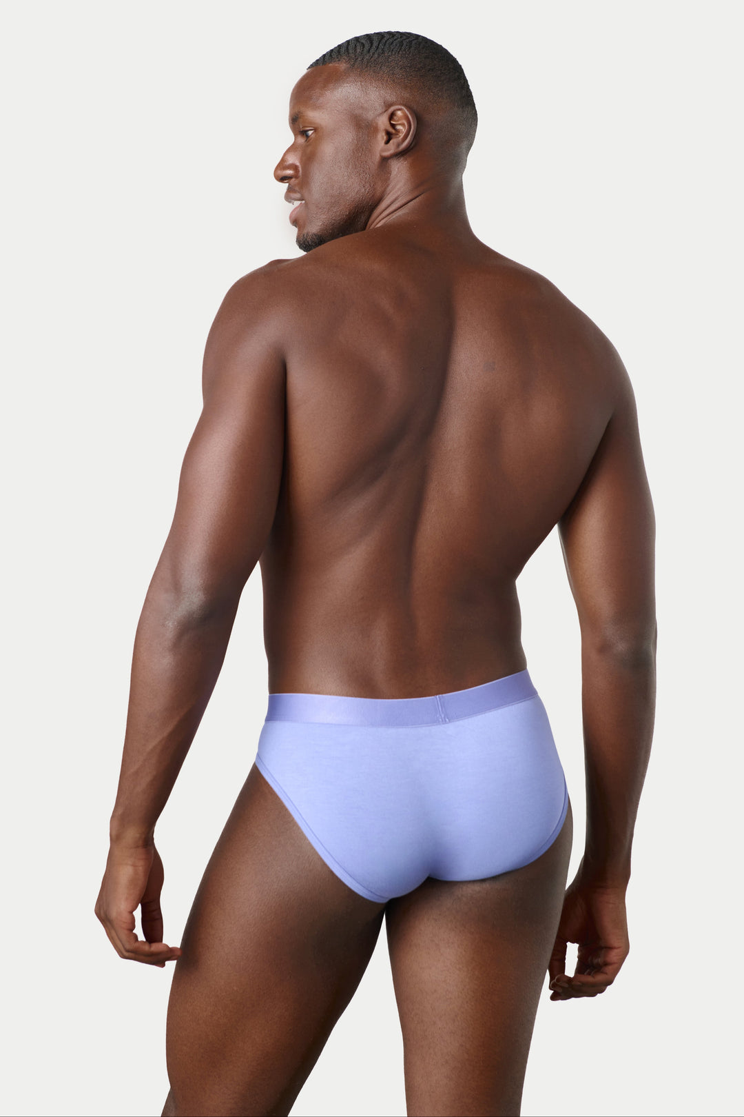 A man shown from the back wearing light purple/lavender briefs, photographed against a clean light gray studio background.