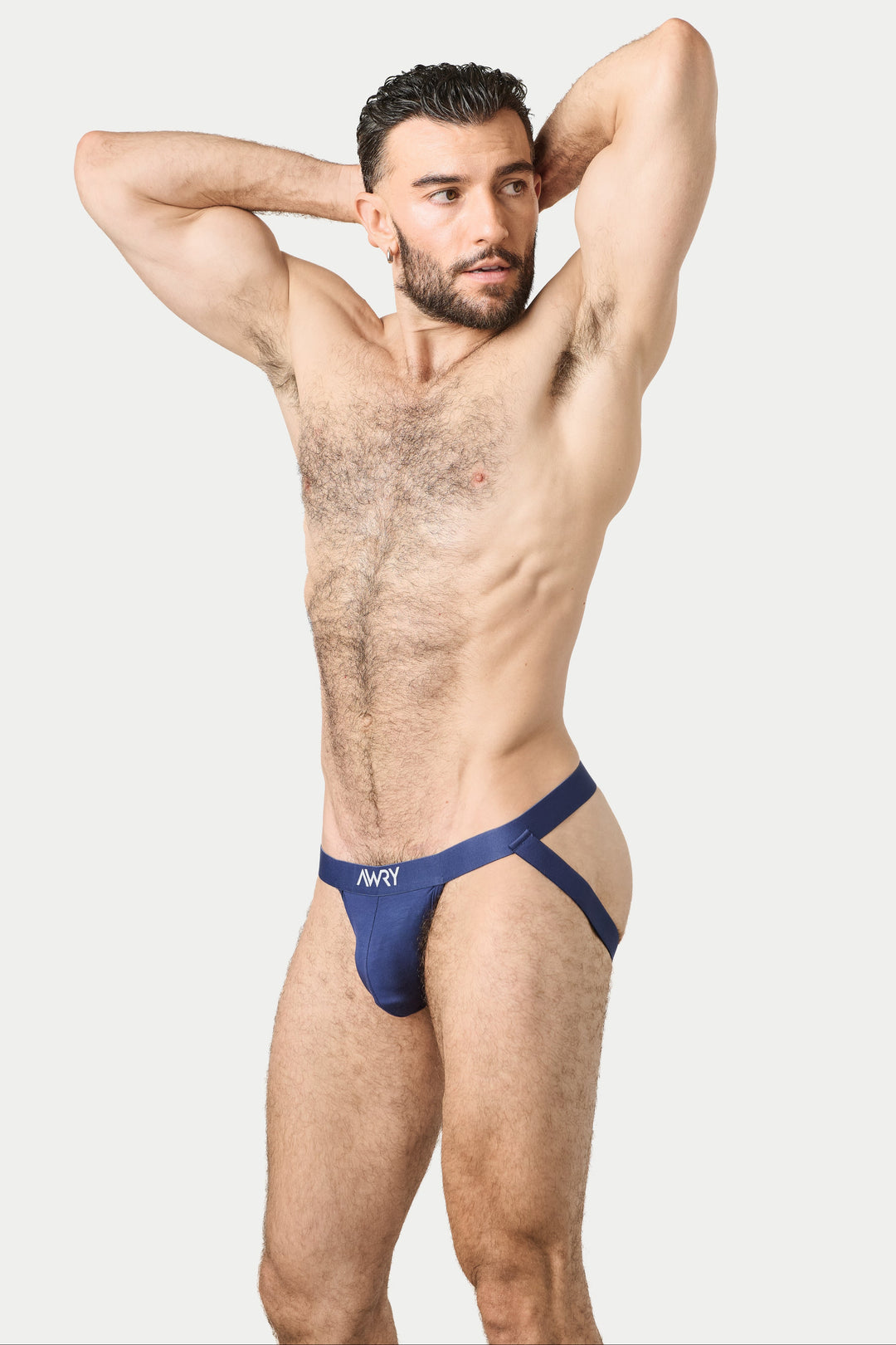 A muscular man posing in a navy blue jockstrap with 'AWRY' printed on the front, set against a solid light gray background.