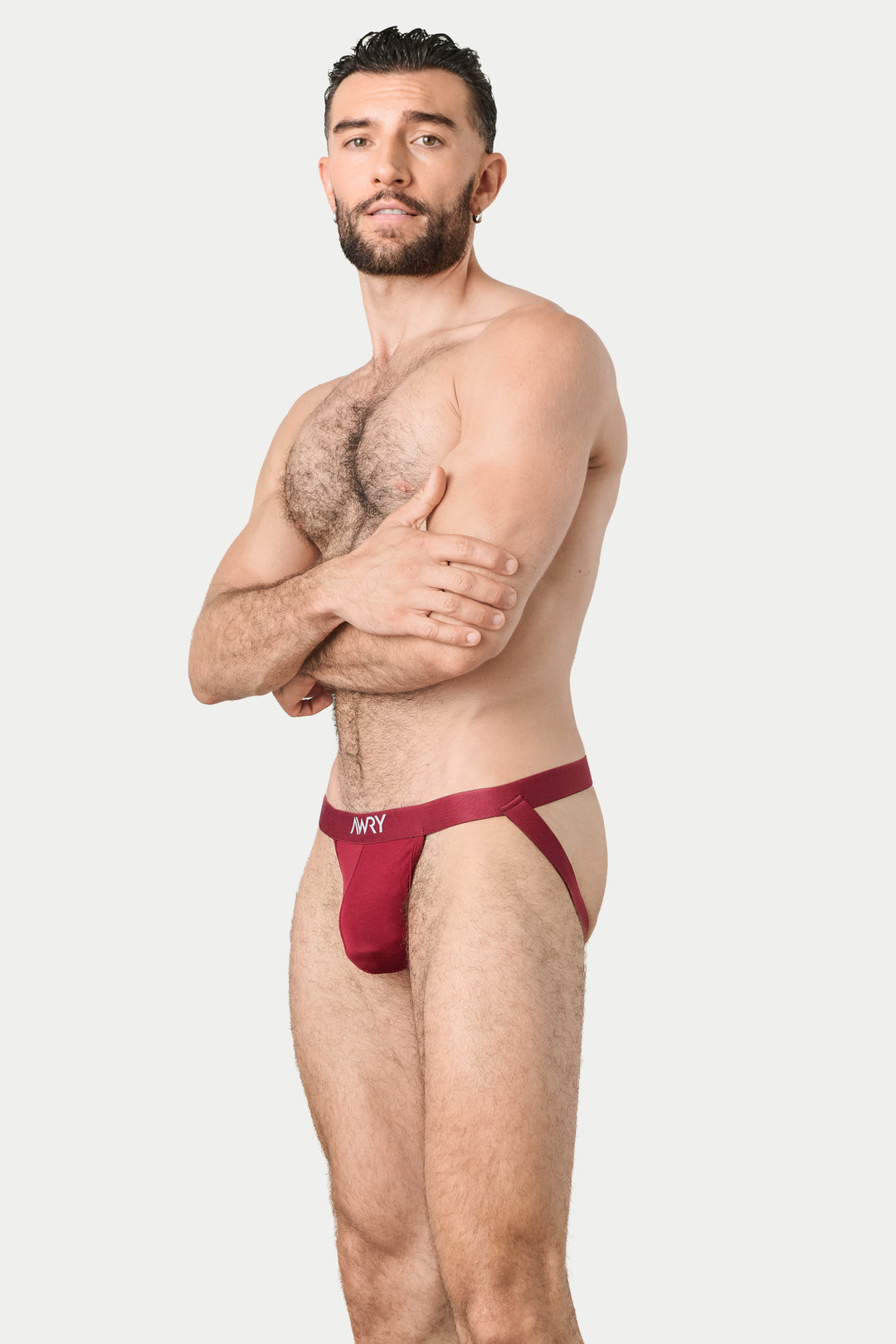 A man modeling a pair of low-rise, jockstrap in a deep burgundy red. The briefs feature a wide waistband with a small white logo that reads 'AWRY'.