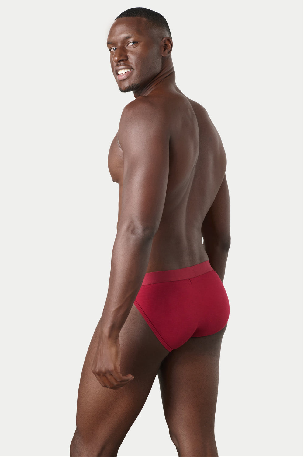 A man wearing red briefs, shown from a side-back angle against a plain light gray studio background.