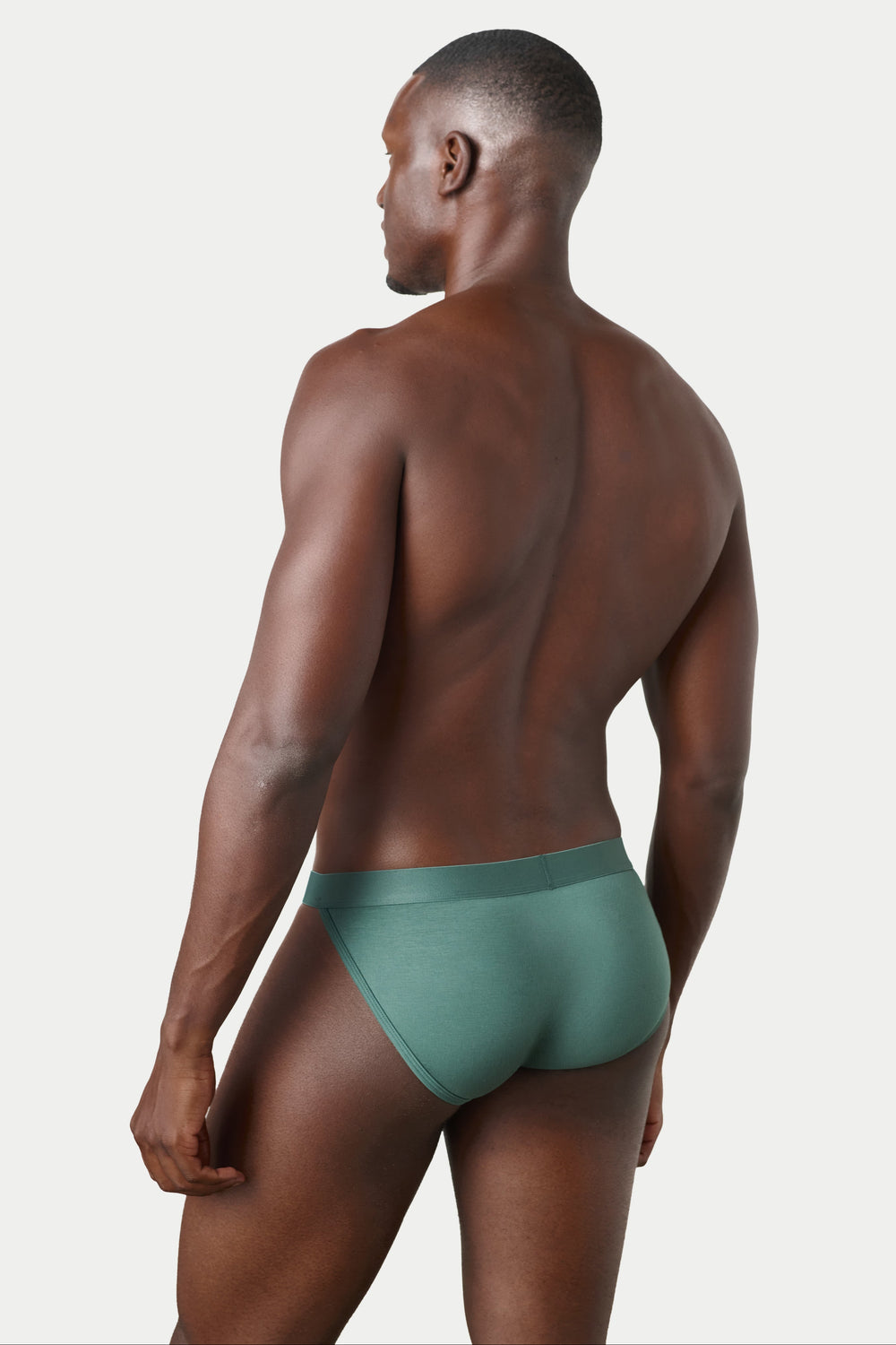 A man shown from the back wearing a pair of solid sage green briefs, photographed against a clean, light gray studio background.