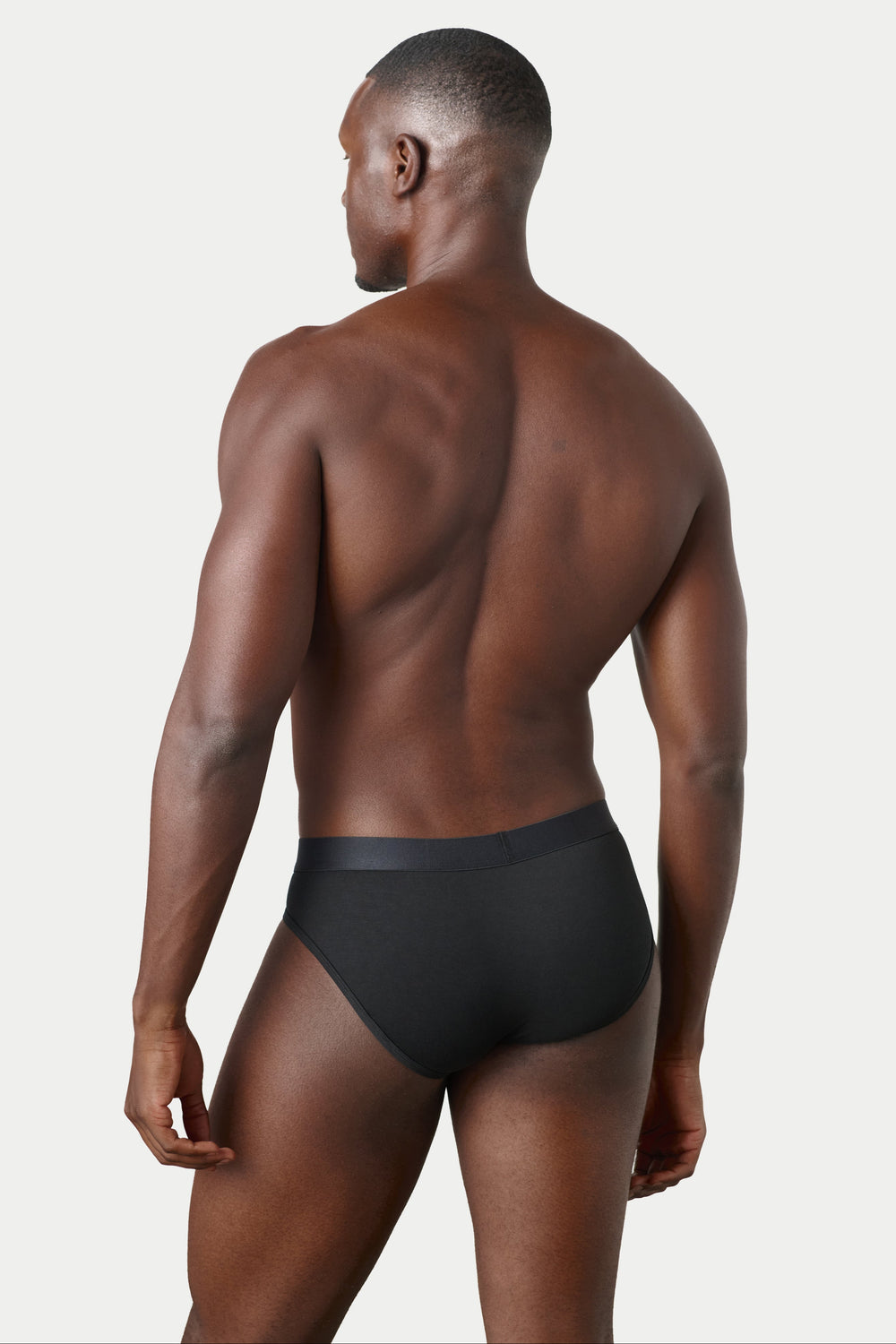 A muscular man shown from the back wearing black briefs, photographed against a clean light gray studio background.