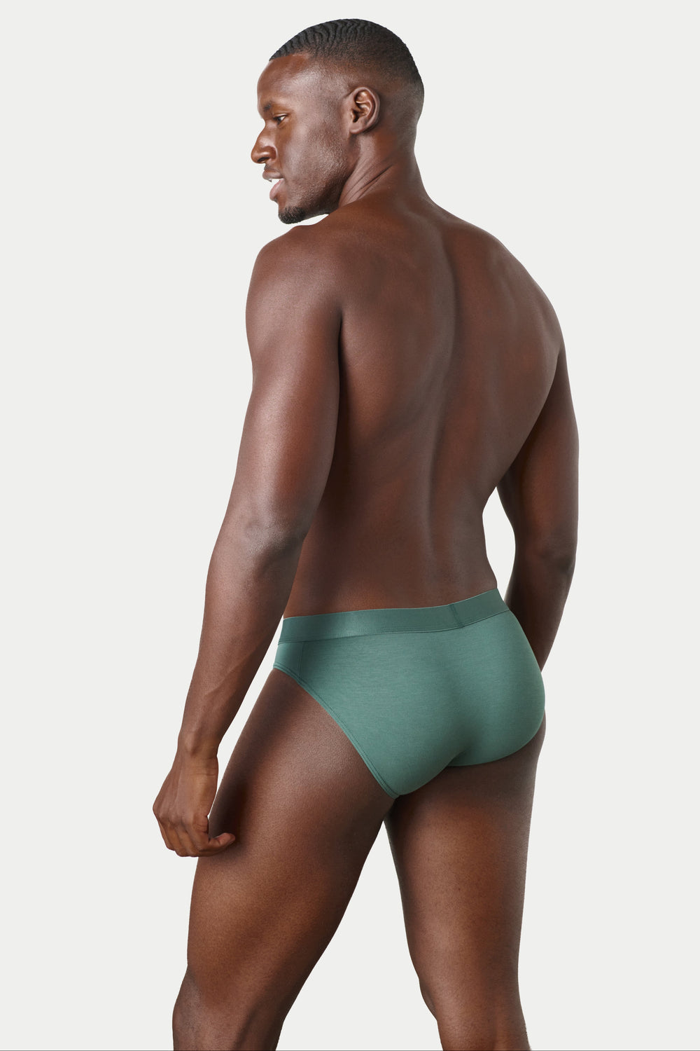 A man shown from the back wearing a pair of low-rise, solid sage green briefs against a light gray studio background.