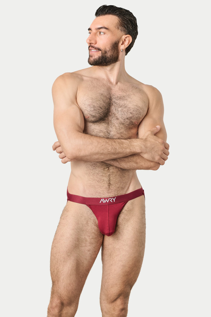 A muscular man with a beard modeling a low-rise, jockthong-style jockstrap in a deep burgundy red. The brand name &
