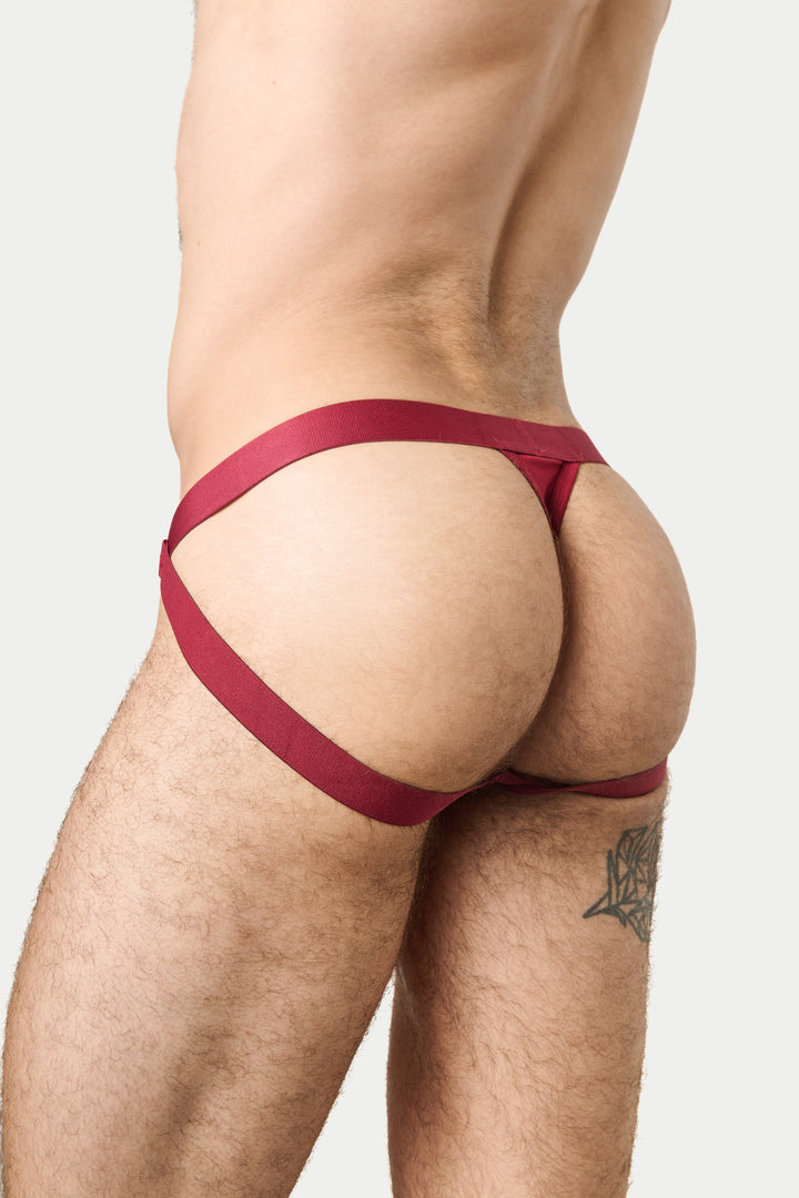 Rear view of a man wearing a dark red jockthong-style brief with wide waist straps, photographed against a light gray studio background.