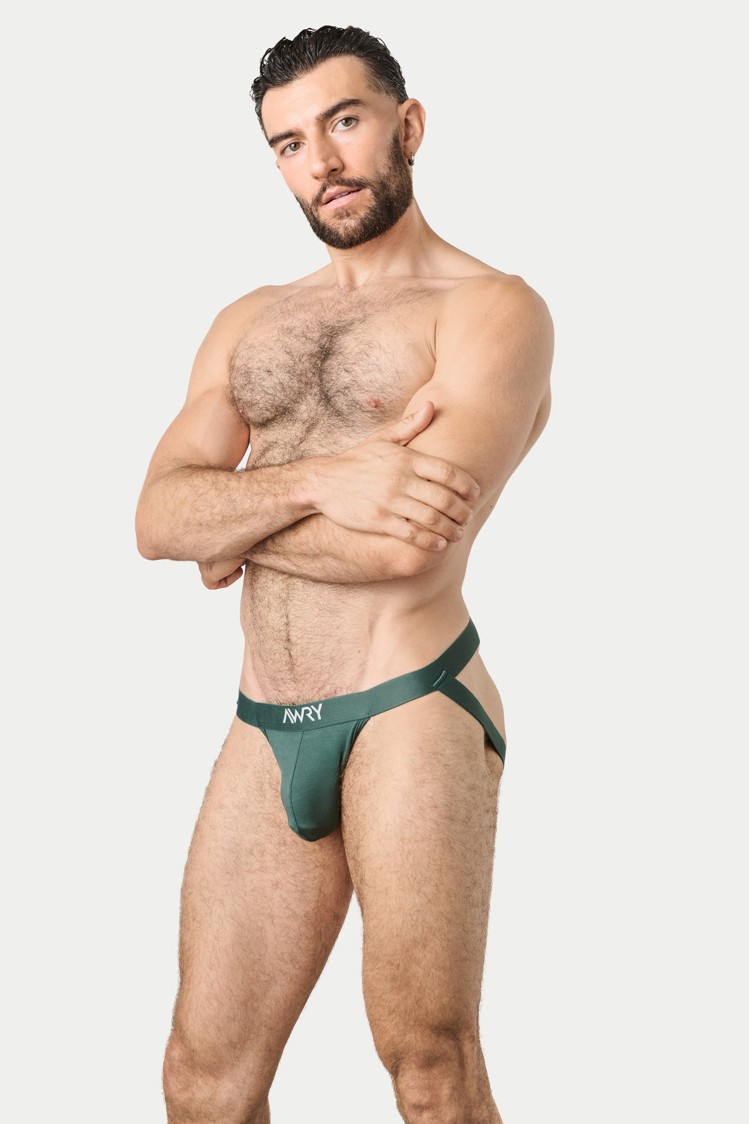 A muscular man with a beard modeling a dark green men's jockstrap with a white 'AWRY' logo on the waistband, posed against a solid light gray background.