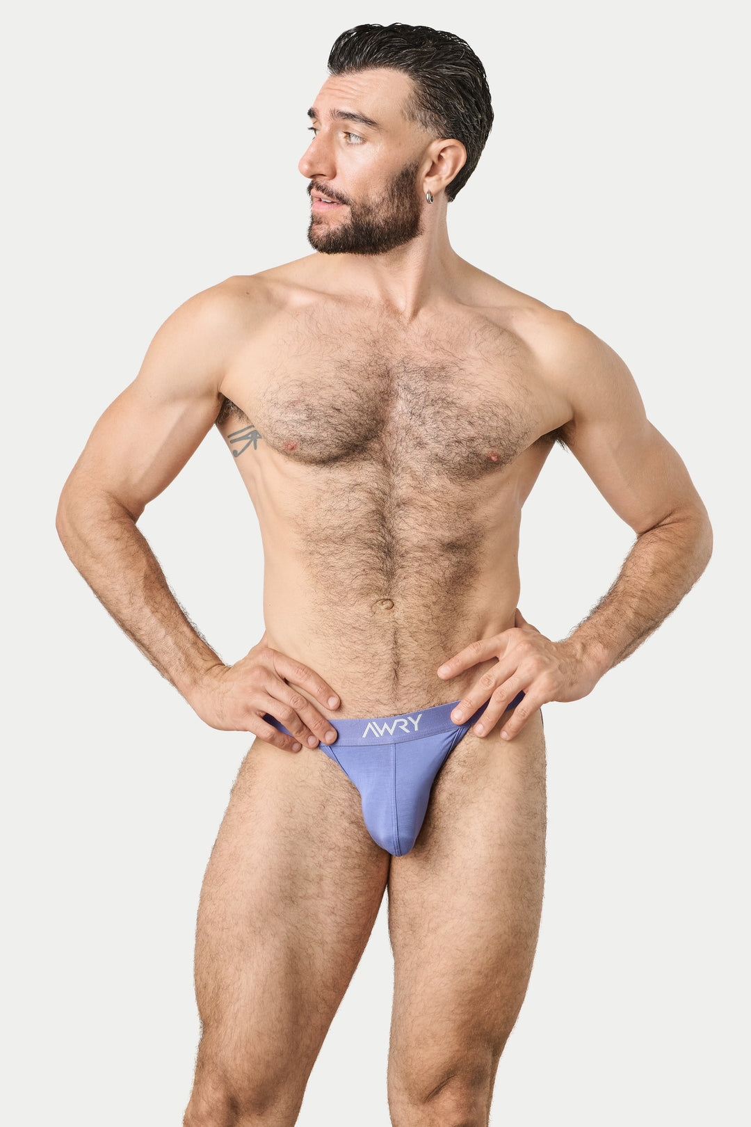 A muscular man with a beard modeling a light purple men's jockthong with a white logo, standing against a solid light gray background.