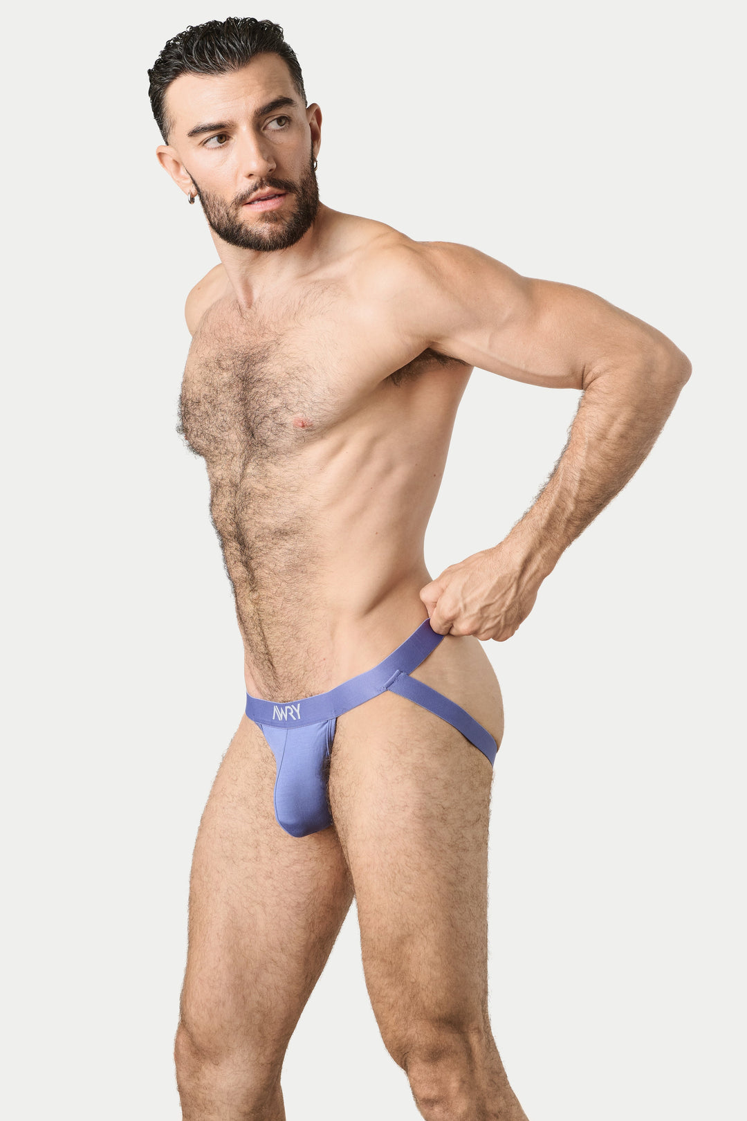 A muscular man modeling a light purple jockthong-style brief with a matching waist strap, posed against a solid light gray background.