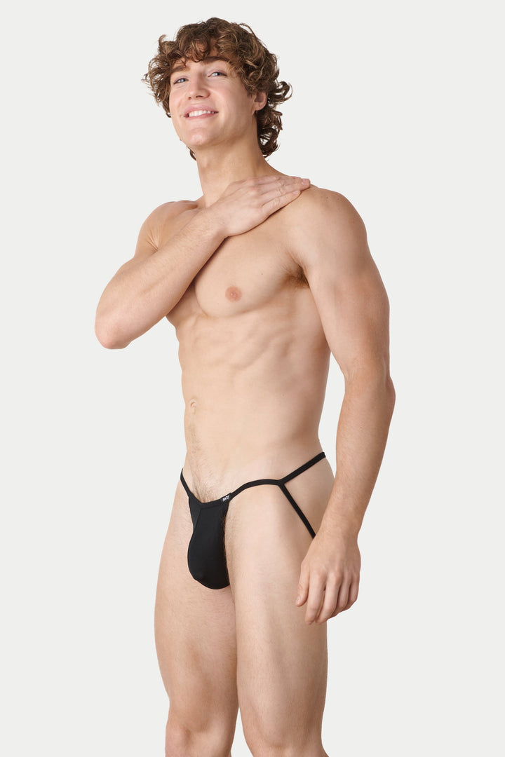 A shirtless male model wearing a minimal black thong-style brief with side straps, standing against a light gray studio background.