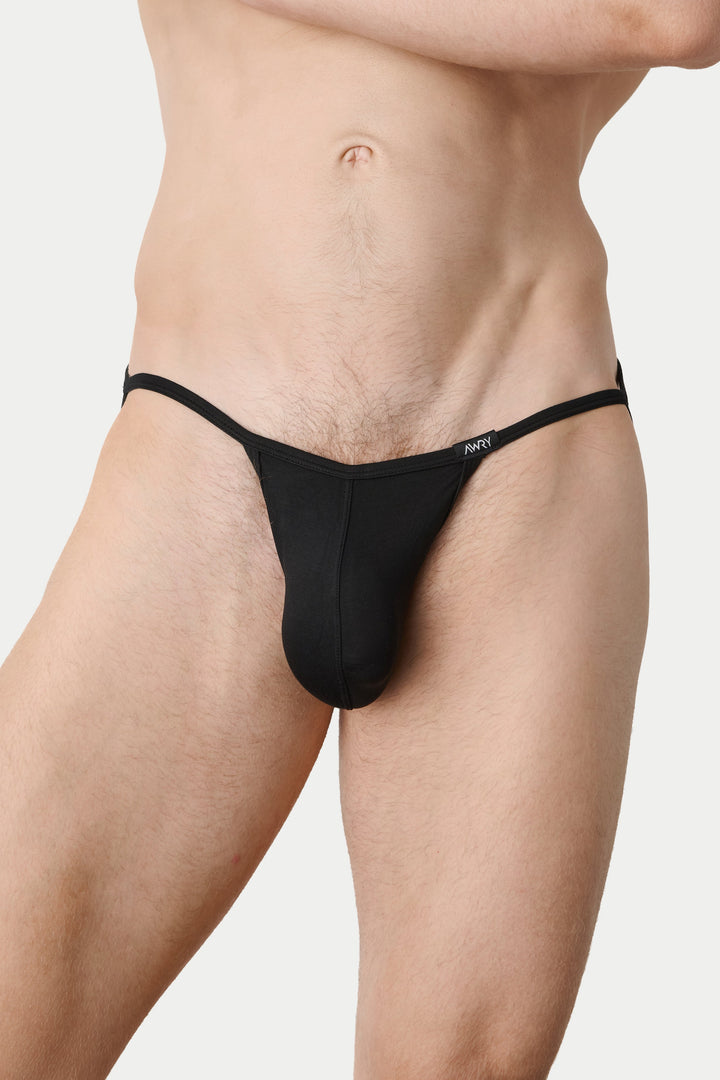 A close-up of a male model wearing a black minimal thong-style underwear brief against a light gray studio background.