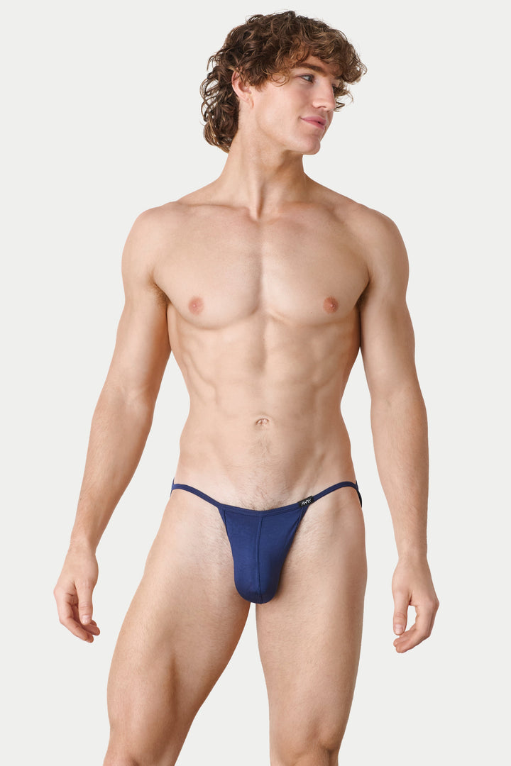 A shirtless male model wearing a minimal navy blue thong-style brief, standing against a plain light gray studio background.