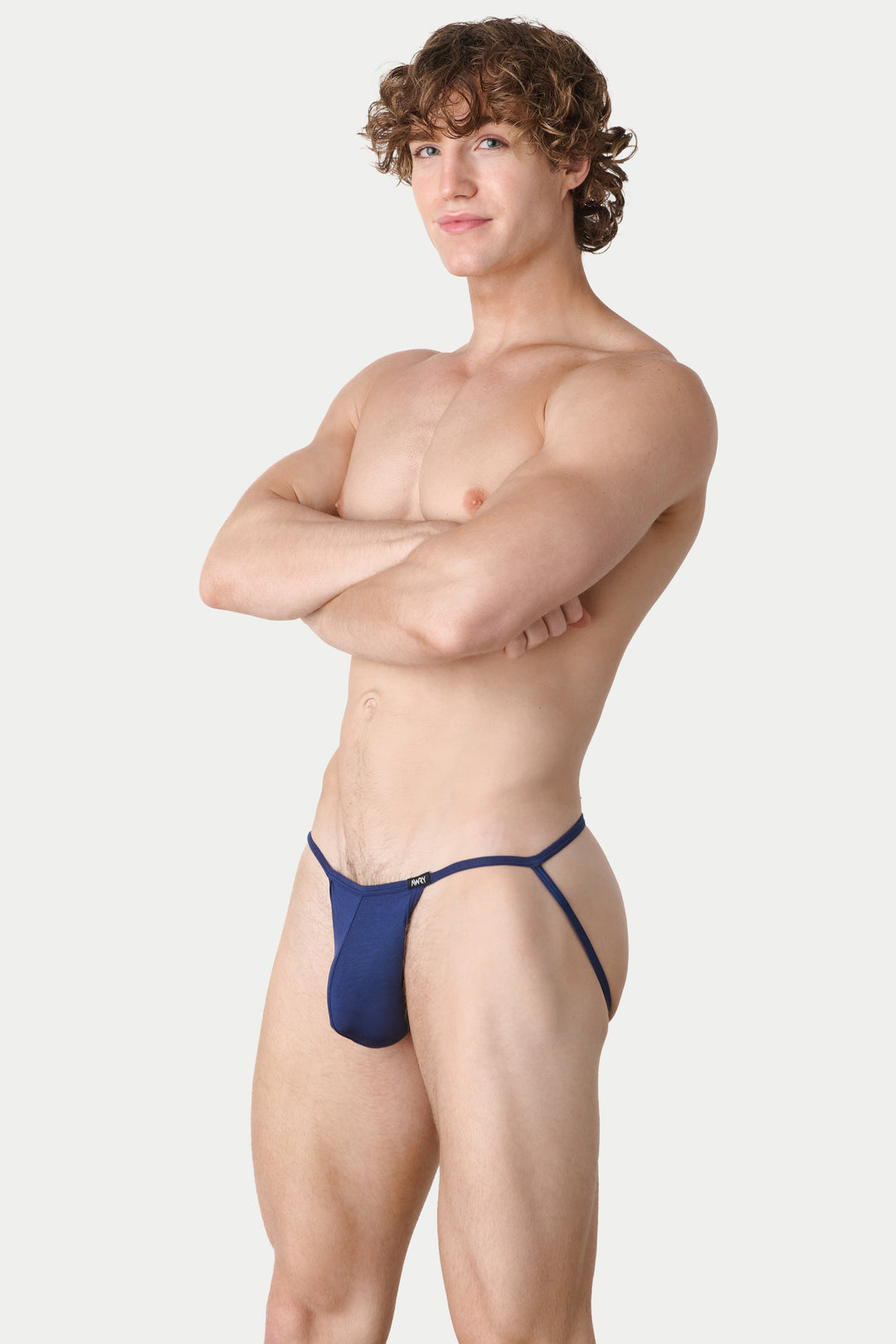 A muscular man with curly brown hair stands with arms crossed, wearing a dark blue thong-style brief against a light gray studio background.