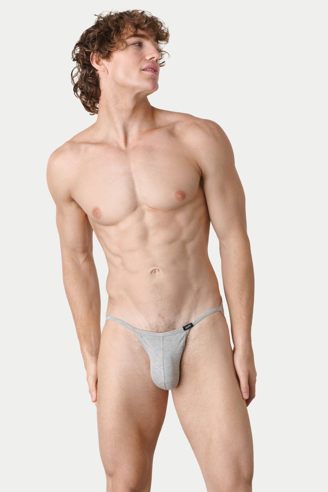 A male model wearing a light gray thong-style brief, standing against a plain light gray studio background.