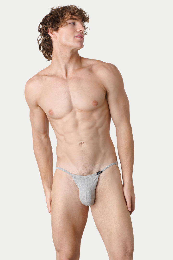 A male model wearing a light gray thong-style brief, standing against a plain light gray studio background.