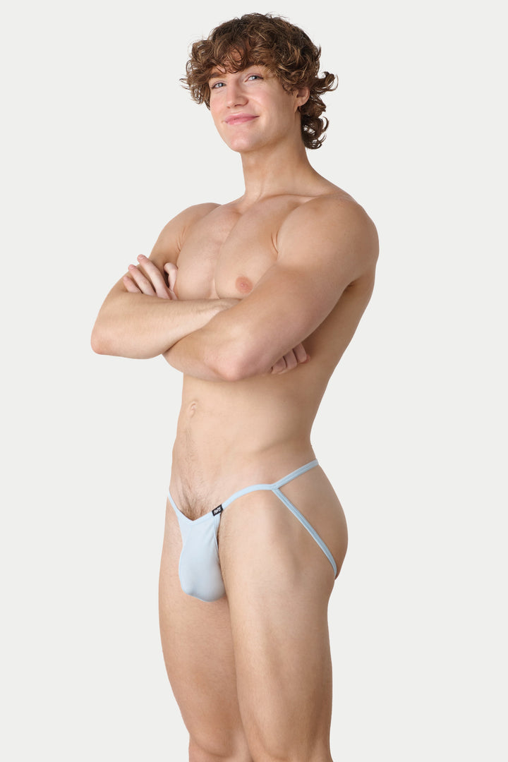 A muscular man with curly brown hair stands in profile against a light gray studio background, wearing a very minimal light blue thong-style underwear piece with thin straps.