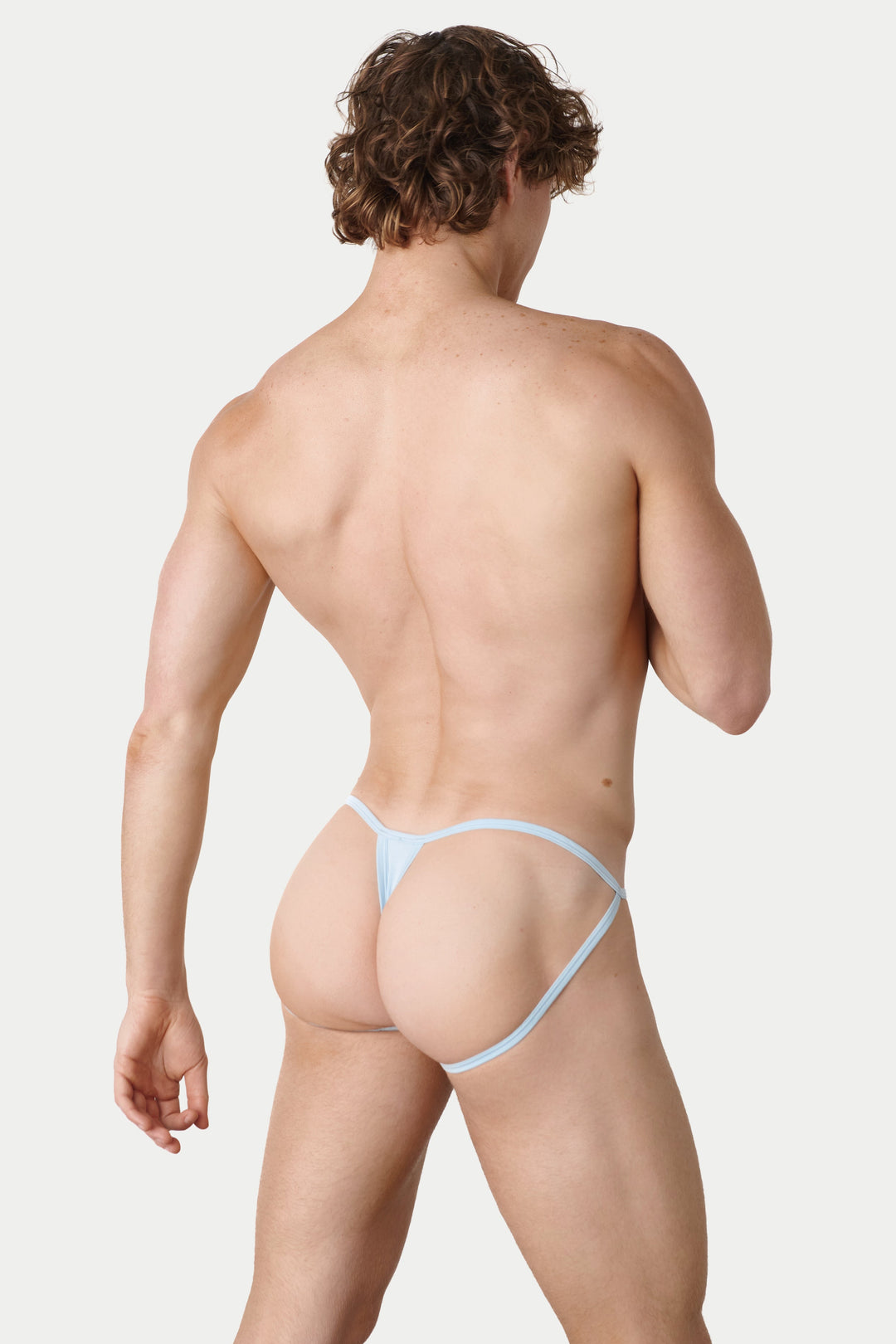 A muscular man with curly brown hair is shown from behind wearing a very minimal light blue thong, against a solid light gray background.