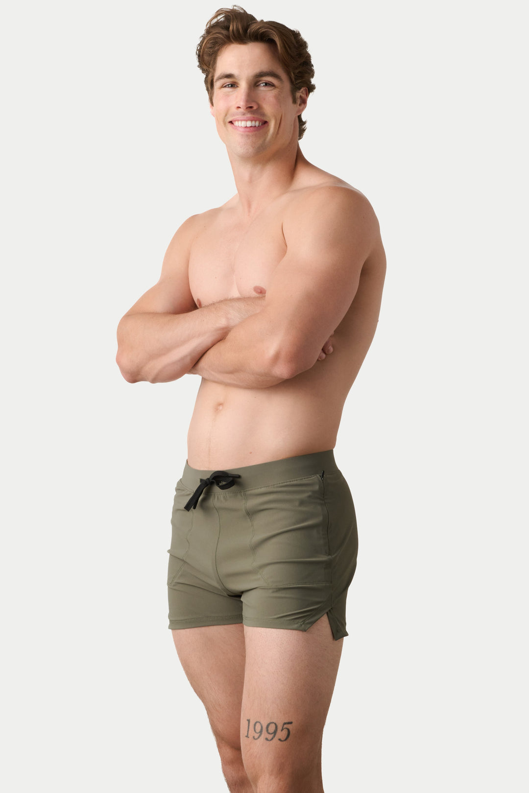 A shirtless man with arms crossed stands against a light gray studio background wearing olive green swim trunks with a black drawstring.