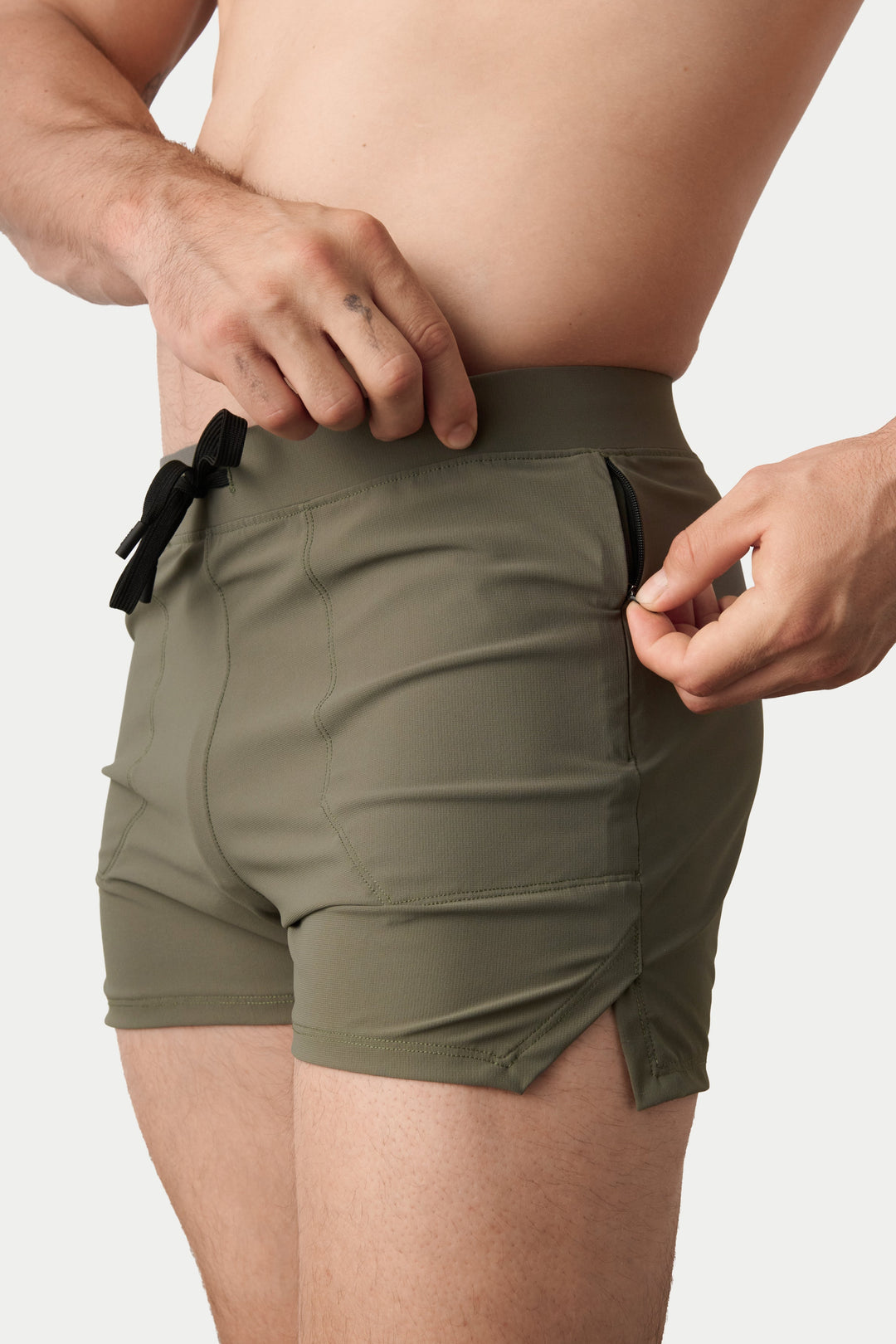 A close-up of a model wearing olive green swim trunks, showing the side pockets and drawstring waistband against a light gray background.