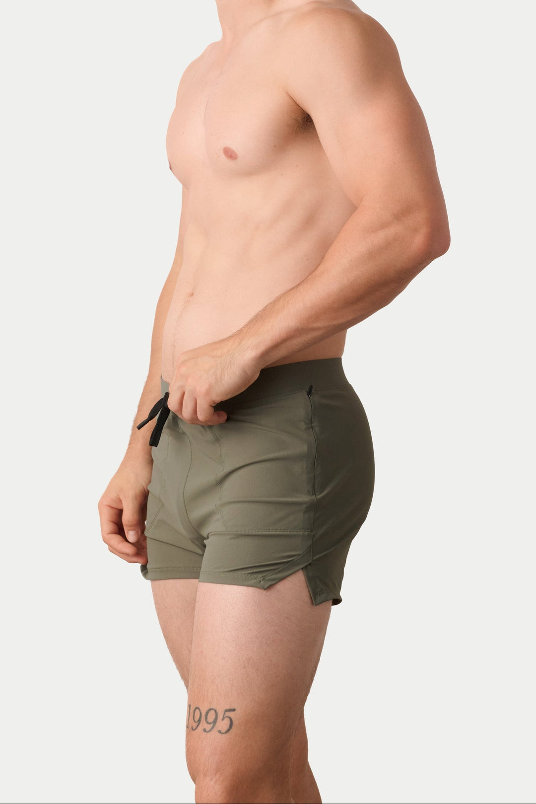 A shirtless man in side profile wearing olive green athletic shorts, standing against a light gray studio background.