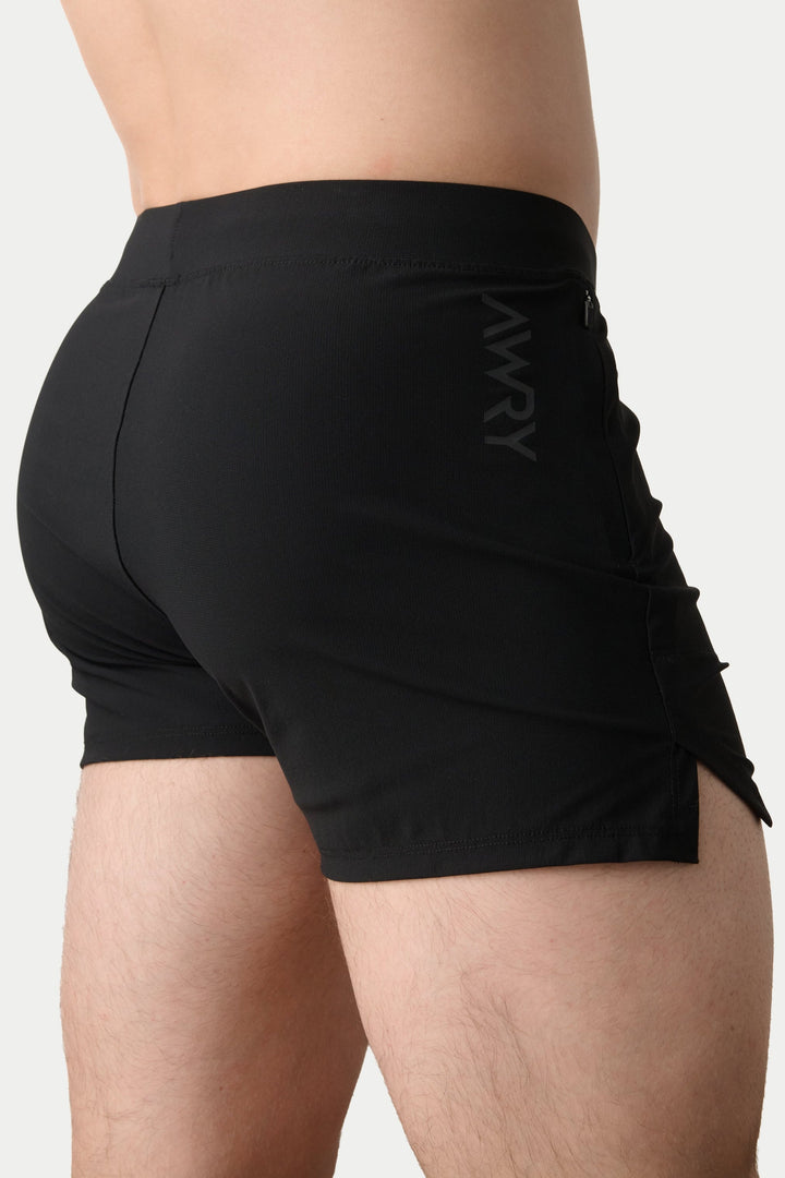 Rear three-quarter view of a model wearing black athletic shorts with a side zipper and a subtle vertical logo, set against a plain light gray background.