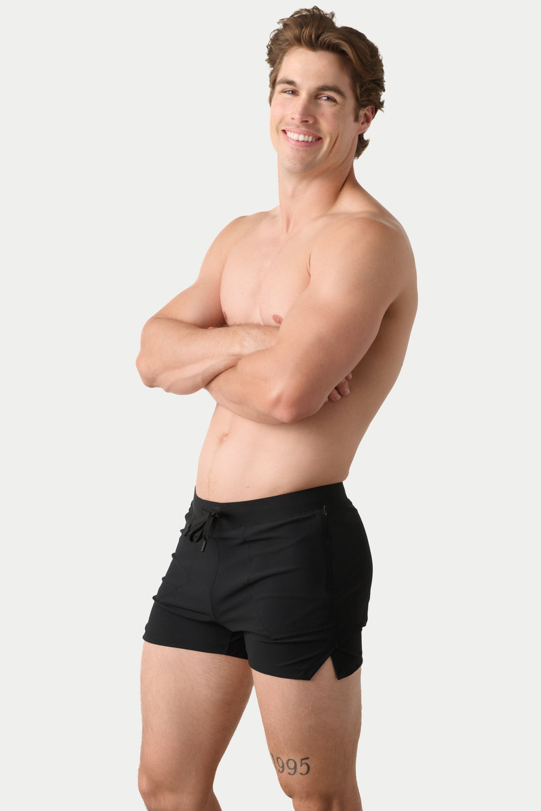 A shirtless man with arms crossed modeling black swim trunks against a light gray studio background.