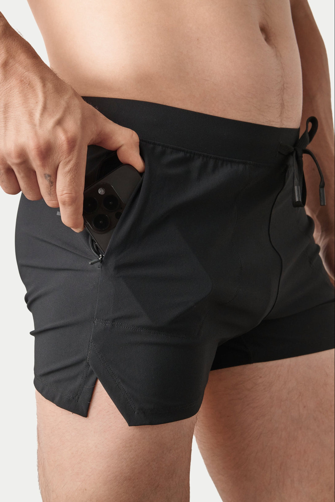 Close-up of a person wearing black athletic shorts with a side zipper and drawstring waist, shown against a light gray studio background.