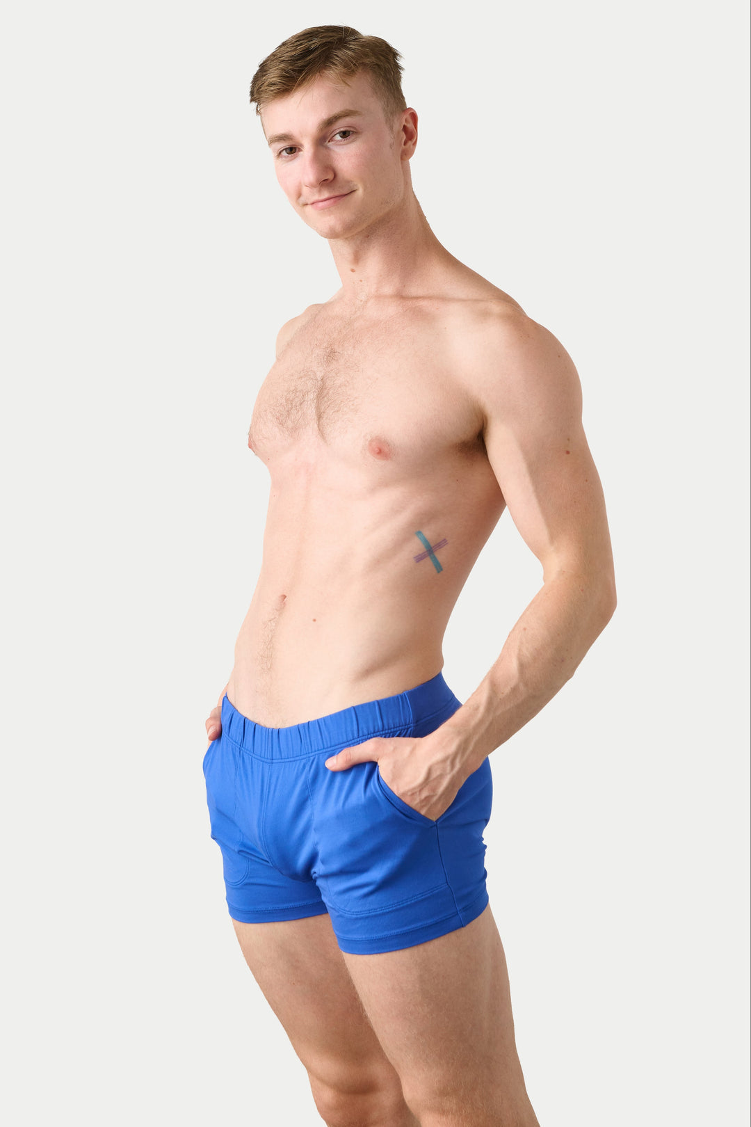 A shirtless man wearing bright blue boxer briefs, standing against a light gray studio background.