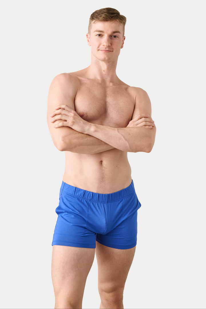 A shirtless young man with arms crossed stands against a light gray studio background, wearing bright blue boxer-brief style shorts.