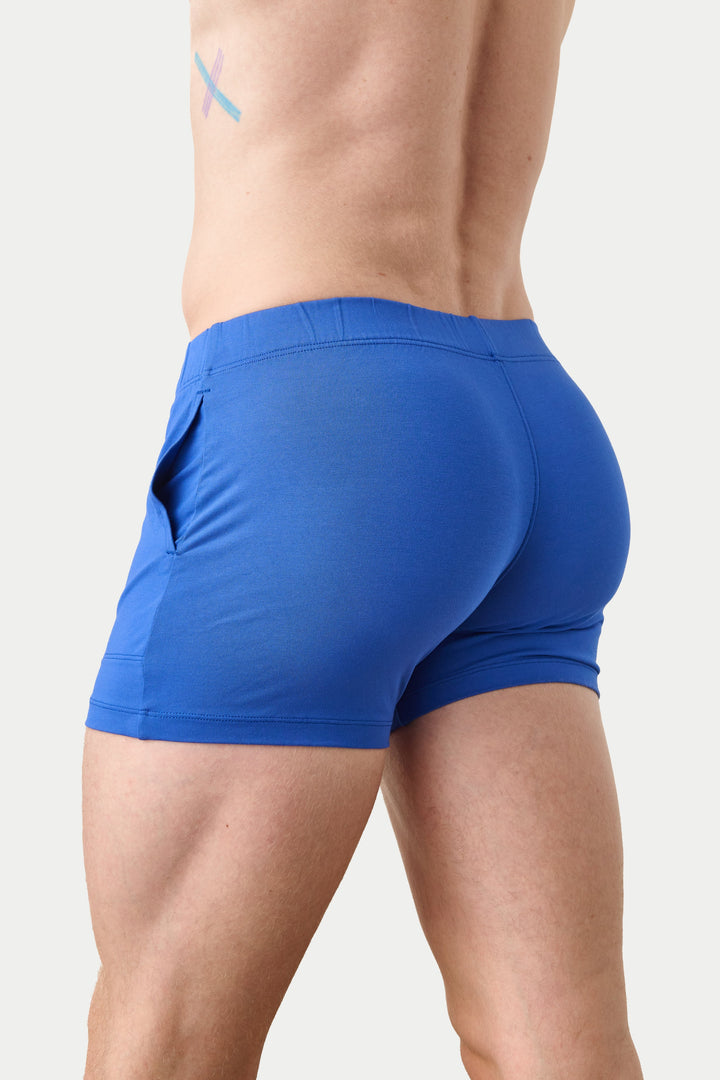 A man wearing bright blue athletic boxer briefs, shown from a rear three-quarter angle against a light gray studio background.