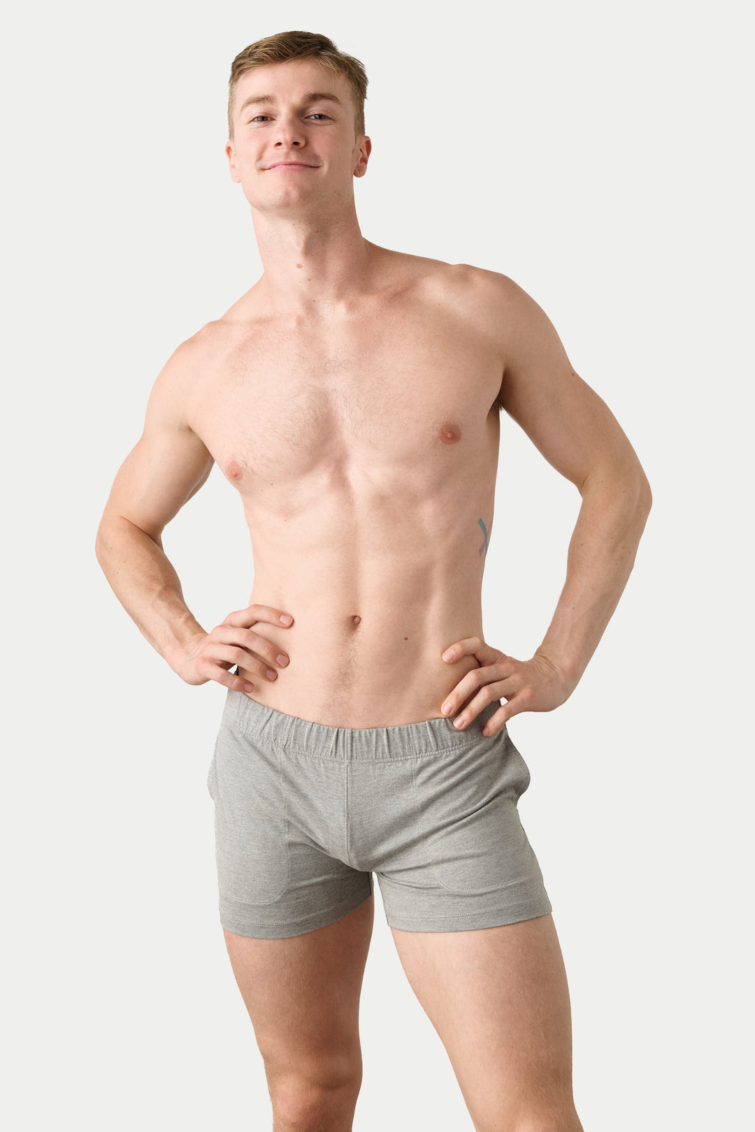A shirtless man modeling light gray boxer briefs against a plain light gray studio background.