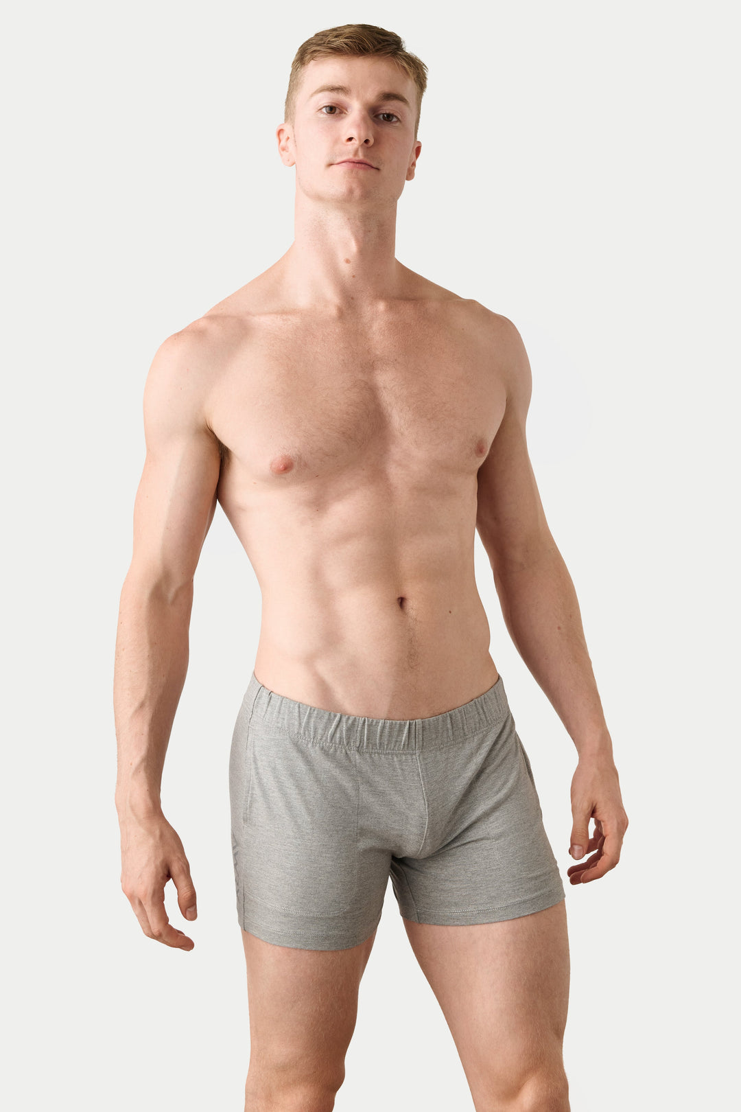 A shirtless male model wearing light gray boxer briefs, standing against a plain light gray studio background.
