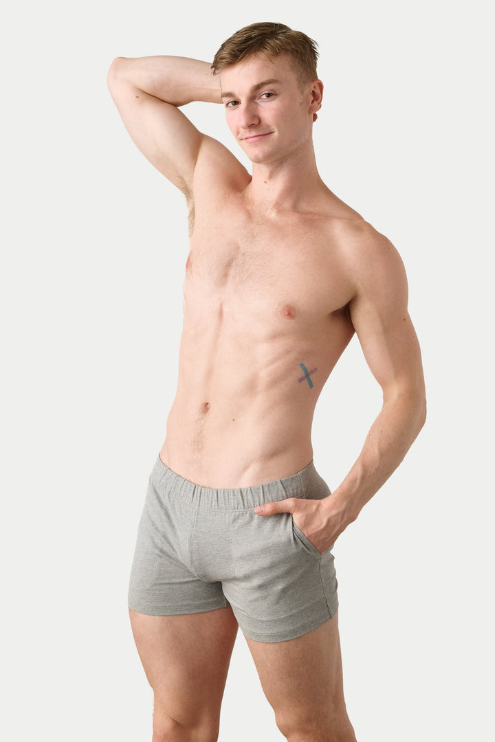 A shirtless young man modeling light gray boxer briefs against a plain light gray studio background.