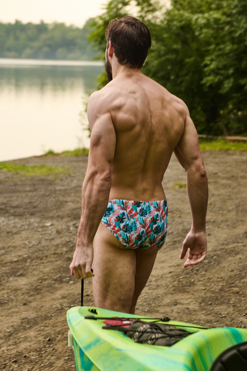 Man wearing tropical print swim brief with multicolored leaf pattern in blue, red, and green tones by a lake.