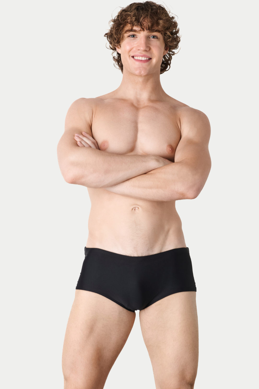 Man wearing black swim briefs with arms crossed against neutral background.