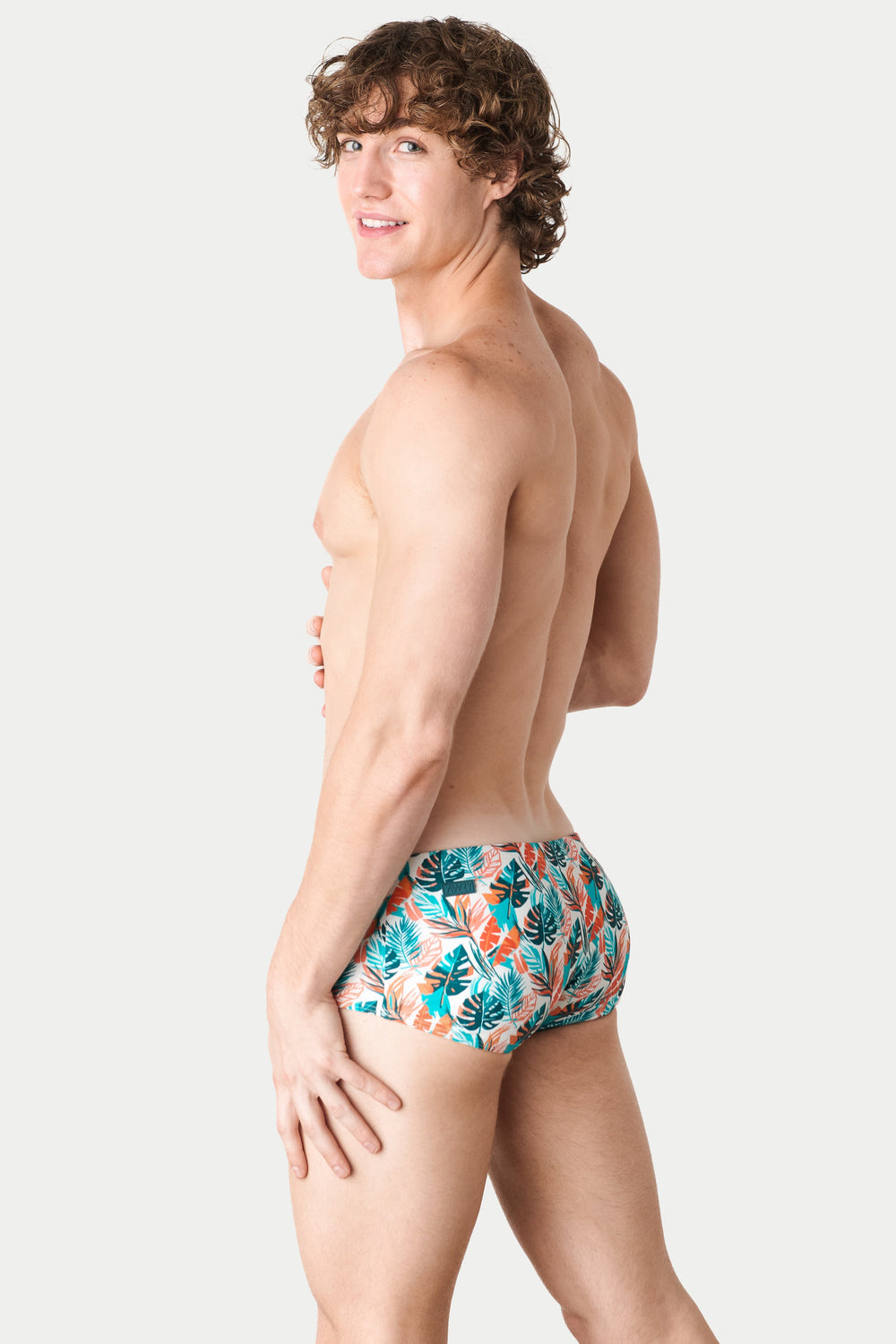 Man wearing tropical print swim shorts with teal, orange, and coral leaf pattern on white background.