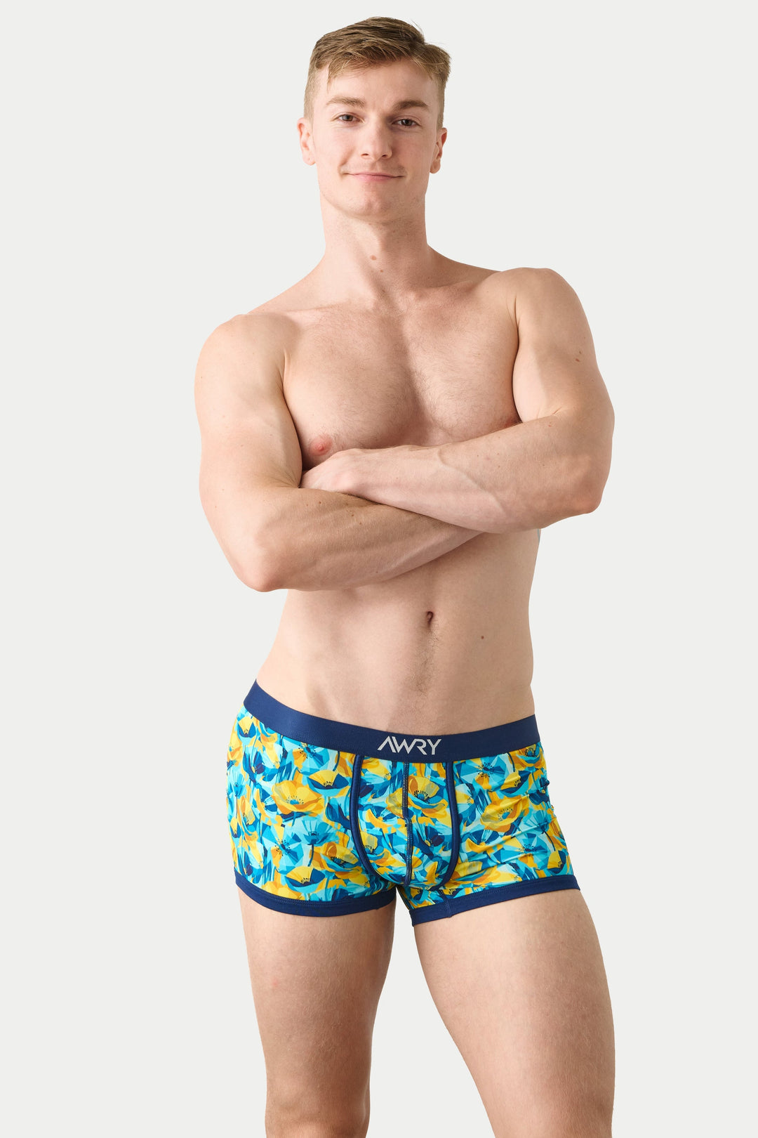 A shirtless male model wearing patterned boxer briefs with a blue waistband and yellow/orange fish print, standing against a light gray studio background.